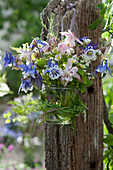 A small bouquet of columbines, wild strawberry and woodruff flowers in a glass jar with a grass wreath hung on a post
