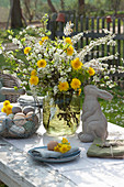 Ostertischdeko im Garten: Strauß aus Blütenzweigen und Löwenzahn, Osterhase, Korb mit Ostereiern und Teller mit Serviette