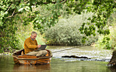 Man using laptop and fly fishing in boat on river