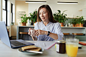 Businesswoman drinking coffee at laptop in morning office