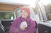 Happy young woman with tea wrapped in blanket at sunny van