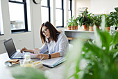 Businesswoman working at laptop in office with plants