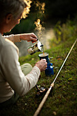 Man with fly fishing pole pouring coffee at riverbank