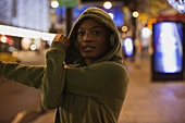 Female runner in hoodie stretching on city sidewalk at night