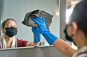 Hotel maid in face mask and glove cleaning bathroom mirror