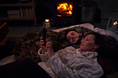 Couple using smartphone on blankets at cozy fireside