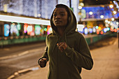 Young woman in hoody jogging on street with lights at night