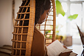 Young woman with headphones reading book in rattan chair