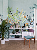 Gepolsterter Stuhl und weißes Regal mit Schubladen vor Tapete mit botanischem Motiv