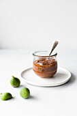 Fig jam jar with a spoon and fresh figs on a white surface.