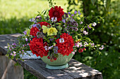 Sommergesteck mit Dahlie 'Garden Wonder', Stockrose, Kronwicke, Dost, Glockenblumen und Himbeeren in Küchensieb