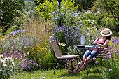 A cozy seating area by the perennial bed with Anise hyssop 'Blue Fortune' 'Apache Sunset', Sea holly 'Glitter Blue', Golden marguerite, grasses, echinacea, petunia, Allium, cape leadwort, and spider flower, glasses, pitcher, and bouquet on the table with a woman relaxing