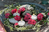 Schale mit Nelkenblüten, Labkraut, Schwimmkerzen und groben Kieselsteinen im Wasser