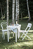 Weißer Tisch mit Geschirr, Kerze und zwei Stühle vor Birken im Frühlingsgarten