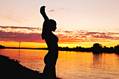 Silhouette of a woman with arms up at riverbank sunset