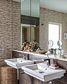 Two washbasins, a shelf and mirrors in a bathroom with patterned wallpaper