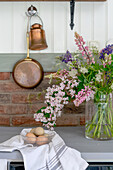 Fresh eggs and summery bouquet of flowers on kitchen worktop below copper pots on wall