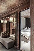Upholstered bench below mirror on wall in elegant, illuminated hallway with wood panelling: view into bedroom
