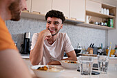 Smiling gay male couple talking and eating in kitchen