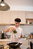 Smiling young man cooking with fresh basil in kitchen