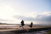 Couple in winter coats running on sunny ocean beach jetty
