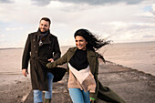 Happy couple in winter coats holding hands on ocean jetty