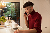 Young man working from home talking on smart phone on laptop