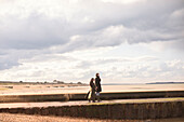 Couple in winter coats walking on sunny winter beach jetty