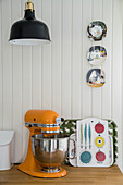 Kitchen appliance and trays below black lampshade and decorative wall plates on wooden panelling