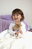 Young girl in pyjamas sitting up in bed, holding toy rabbit