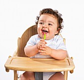 Baby boy in high chair, holding spoon and laughing