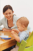 Woman sitting at table, smiling at baby with carrot on plate
