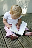 Blonde baby girl sitting on patio looking at picture book