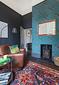 Leather armchairs on colourful rug in front of wood-burning stove; blue wallpaper with antler motif