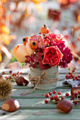 Kleines Herbstgesteck aus Rosen, Hagebutten und Hortensienblüten, Vase mit Stoffband umwickelt