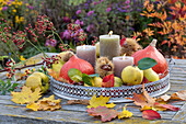 Herbstliche Dekoration mit Hokkaido-Kürbissen, Apfelquitten, Maronen, Hagebutten, Kerzen und buntem Herbstlaub auf Metall-Tablett