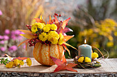 Herbststrauß aus Chrysanthemen, Hagebutten und Herbstlaub in Kürbis als Vase, Kerze im Graskranz mit Blüten dekoriert