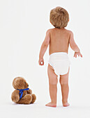 Child in nappy standing beside teddy bear