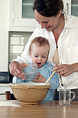 Woman helping baby boy stir wooden spoon in large bowl