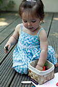 Baby girl sitting on decking picking crayons from a basket