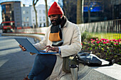 Businessman in stocking cap working at laptop in city park