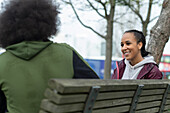 Happy young woman talking with friend on park bench