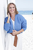 A long-haired woman wearing a white summer dress and a blue cardigan on a beach