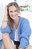 Langhaarige Frau in weißem Sommerkleid und blauer Strickjacke am Strand sitzend