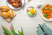 Easter food on white rustic table - pastel colored eggs, roasted chicken and vegetables, buns and spring flowers tulips