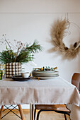 Vorbereitung für einen weihnachtlichen Esstisch: Vase mit Kiefernzweig, Tellerstapel und goldenes Besteck, Floral Hoop an der Wand