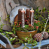 Adventsgesteck mit Hagebutten und braunen Kerzen, umgeben von Zapfen und Mistelzweigen