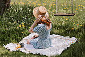 Female in dress and straw hat sitting on picnic blanket on green meadow near swings in summer countryside