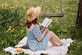 Female in dress and straw hat reading novel while sitting on picnic blanket on green meadow near swings in summer countryside