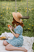 Rothaarige Frau im Kleid und Strohhut sitzt barfuß auf einer Picknickdecke und trinkt